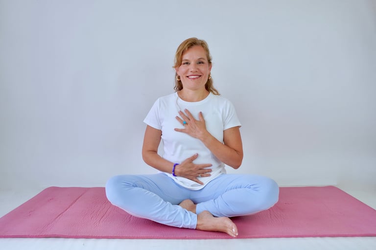 Young girl sitting cross-legged on a pink yoga mat in a white shirt and light blue pants, smiling at the camera