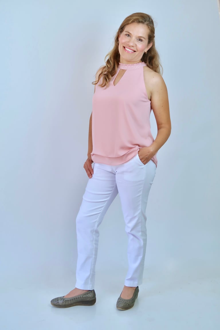 Woman smiling at camera wearing pink sleeveless top with choker detail, white pants, and gray loafers against blue background