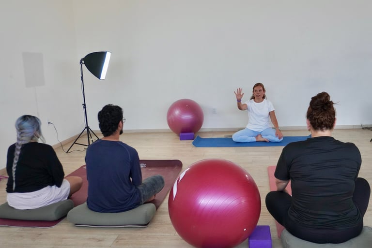 Fitness class with adults and children sitting on yoga mats holding large pink exercise balls in a studio with lighting equipment